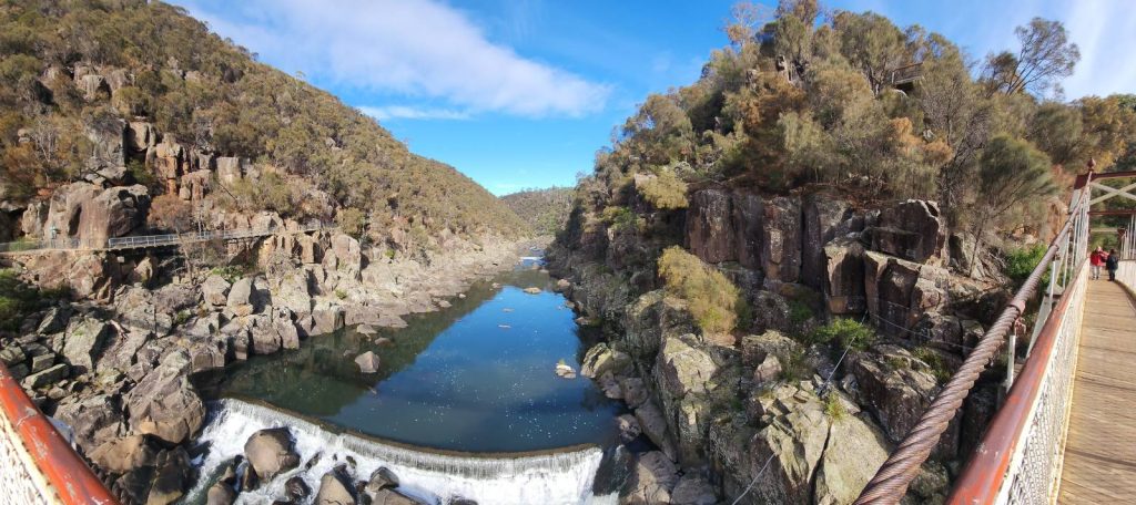 Sunlit rocky river gorge with steep bushland cliffs and a footbridge crossing the water